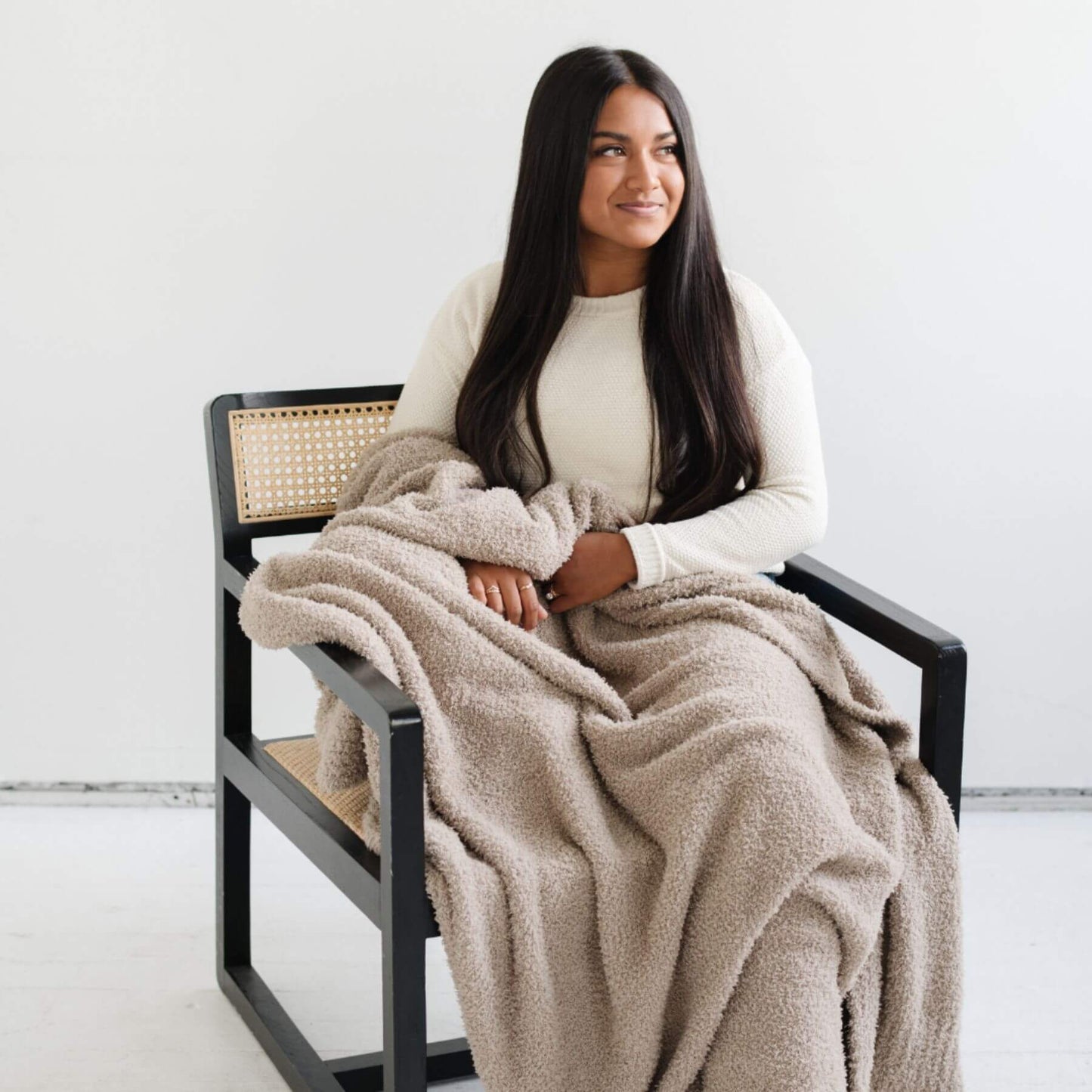 Woman sitting on a chair with Saranoni Bamboni® Throw Blanket draped over her, smiling.