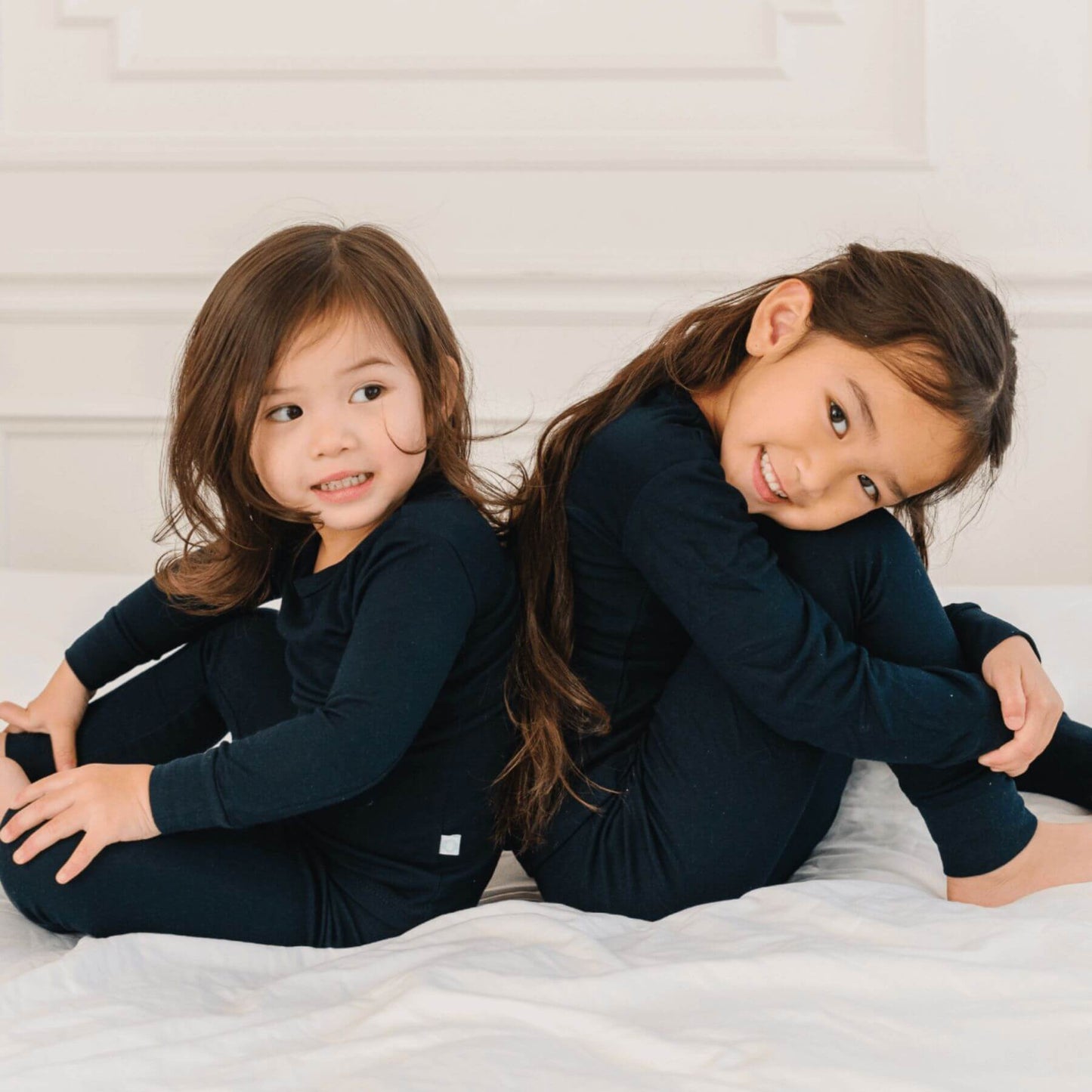 Two young girls in Dreamland Baby Toddler/Kids Bamboo Pajamas - Midnight Black sitting on a white bed.