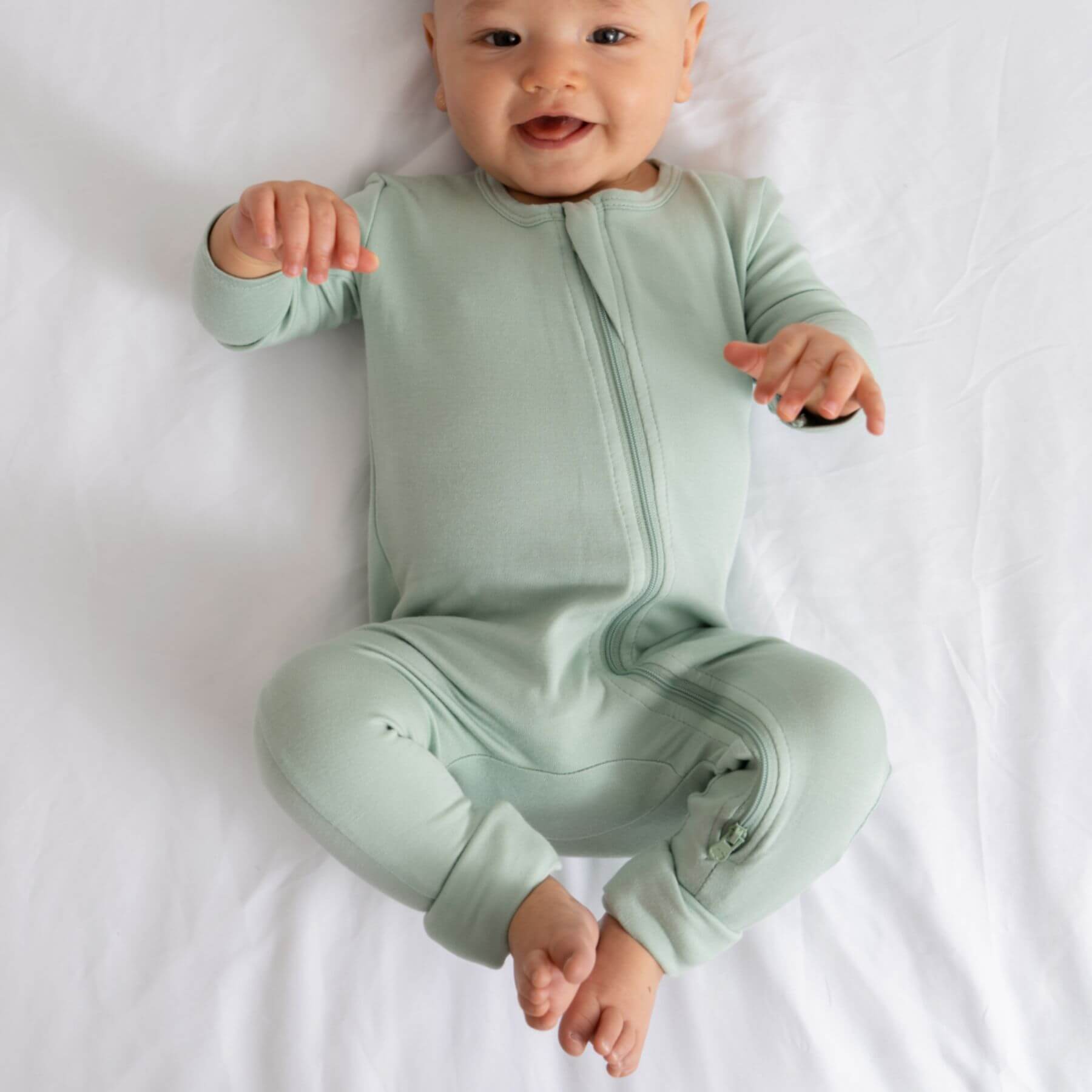 Baby wearing Dreamland Baby Bamboo Pajamas w/ DreamCuffs® - Sage on a white bed