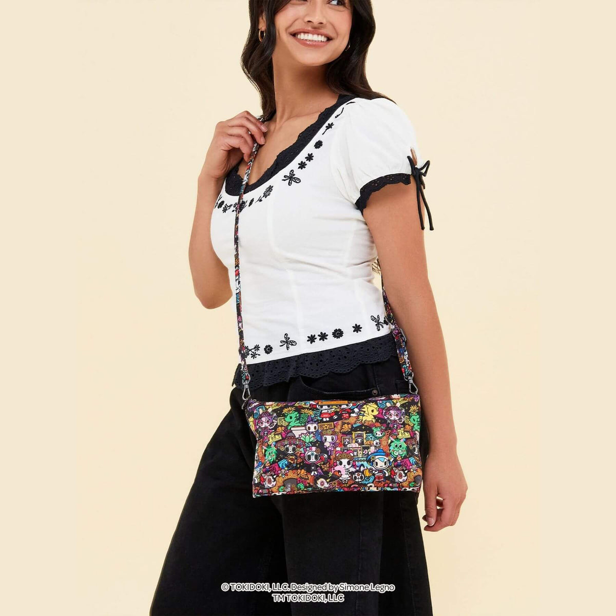 A woman wearing a white short-sleeve top with black lace trim and a black skirt, carrying tokidoki Be Quick Crossbody Diaper Bag Accessory – Dia de los Muertos.