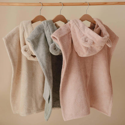 Three Mushie Bear Poncho Towels in beige, gray, and pink hanging on a wooden rod against a beige background.