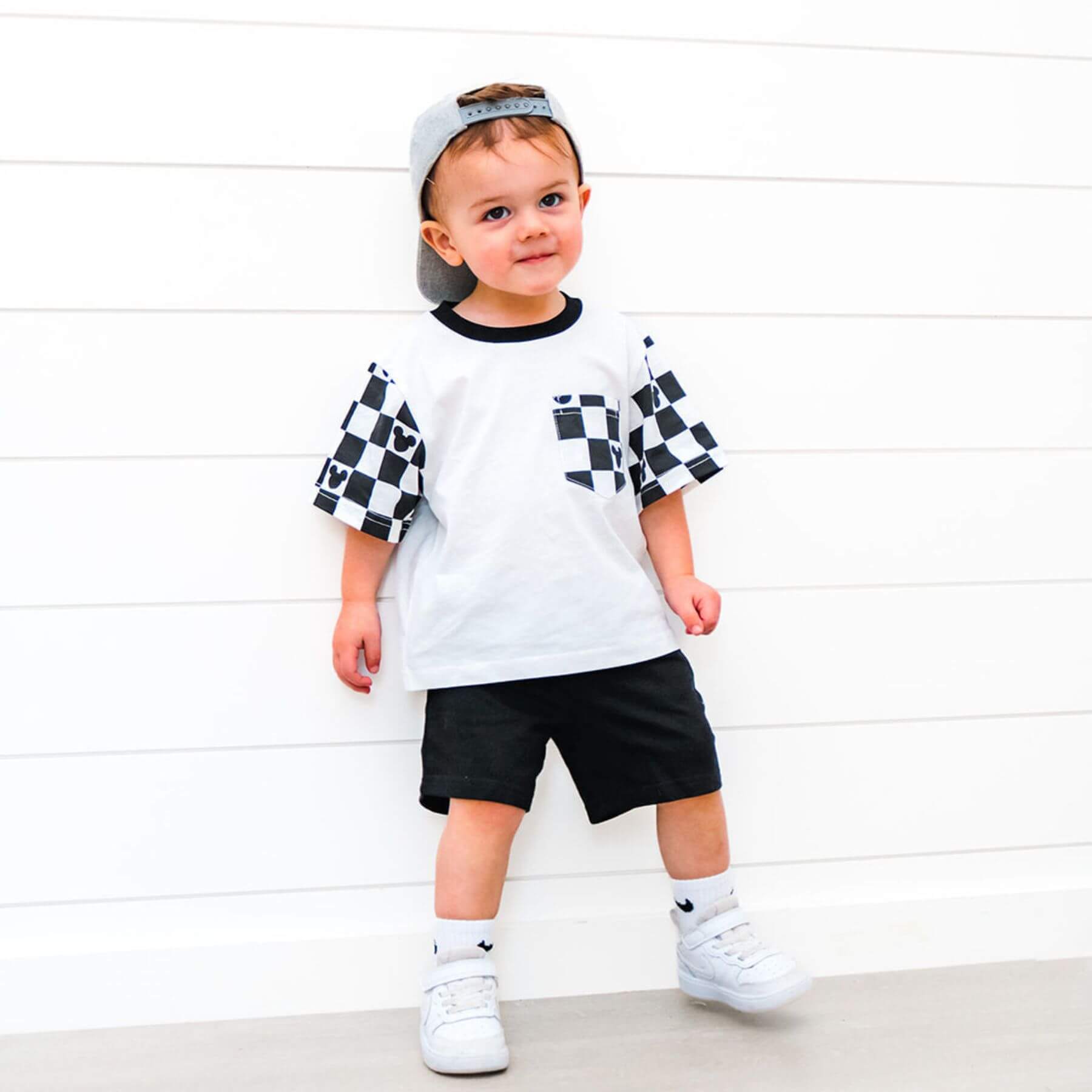 Child wearing Sweet P Baby Co. Black Checkered Pocket Toddler & Kids T Shirt against a white wooden panel background.