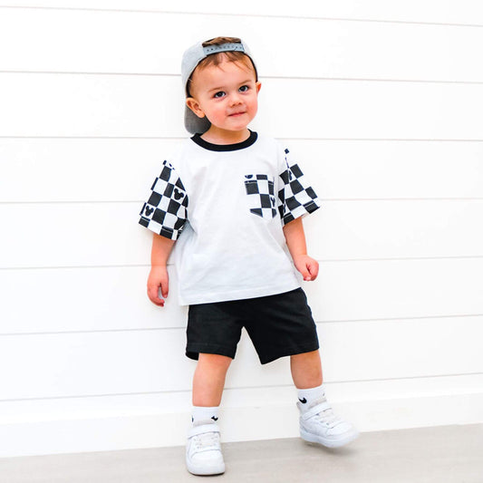Child wearing Sweet P Baby Co. Black Checkered Pocket Toddler & Kids T Shirt against a white wooden panel background.