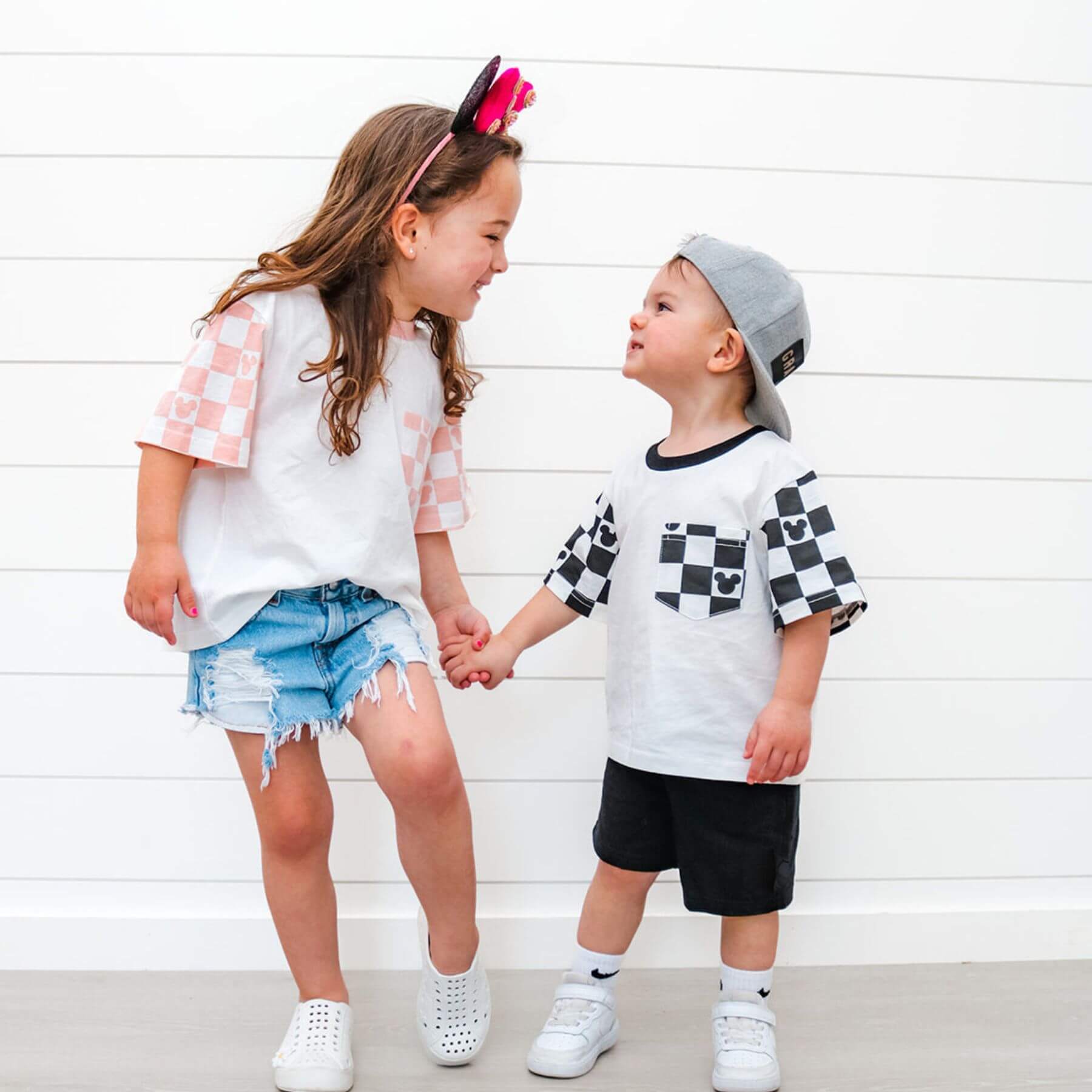 Two children holding hands wearing Sweet P Baby Co. Black Checkered Pocket Toddler & Kids T Shirt against a white wooden background
