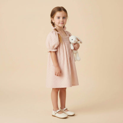 A young girl with braided hair stands against a solid beige background, wearing Makemake Organics Organic Eyelet Bubble Sleeve Dress - Blush Eyelet