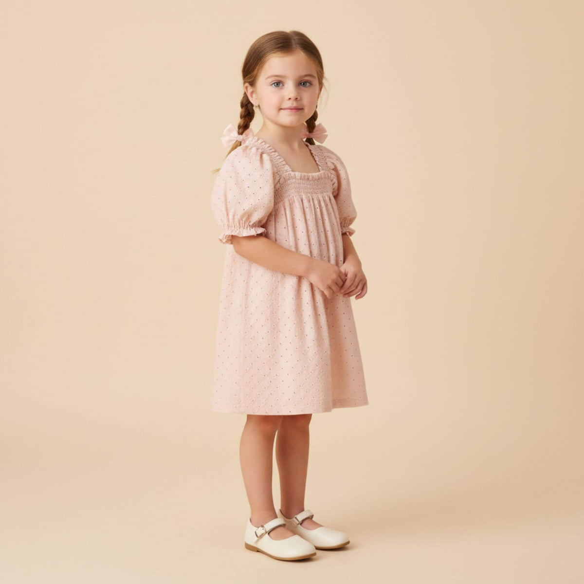 A young girl with braided hair stands against a solid beige studio background wearing Makemake Organics Organic Eyelet Bubble Sleeve Dress - Blush Eyelet
