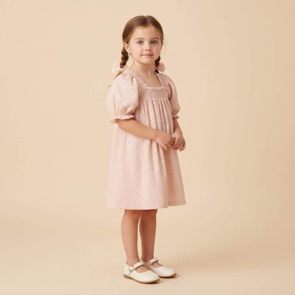 A young girl with braided hair stands against a solid beige studio background wearing Makemake Organics Organic Eyelet Bubble Sleeve Dress - Blush Eyelet