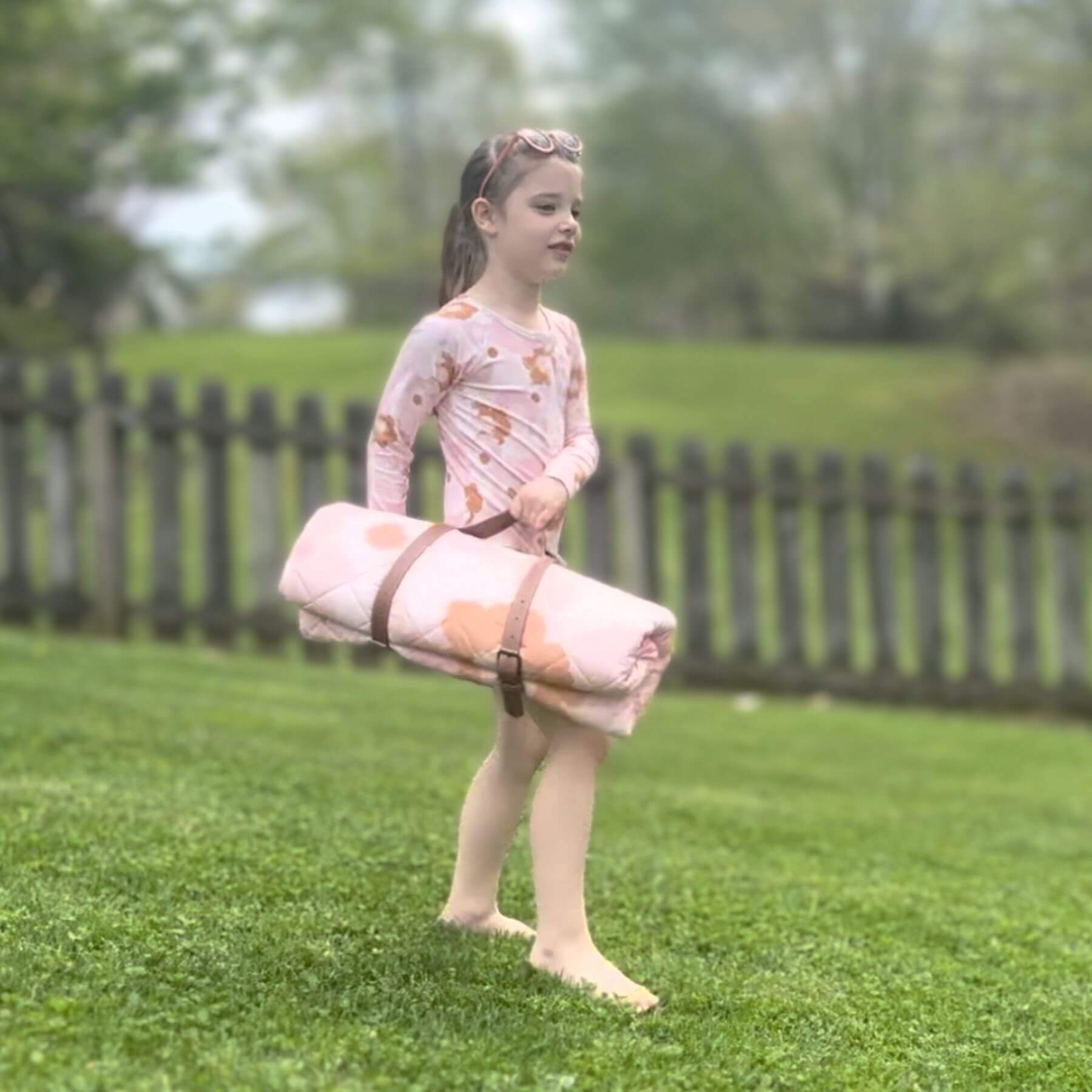 Young girl holding Blush Meadow Quilted Water Resistant Beach Mat on grass with a wooden fence in the background