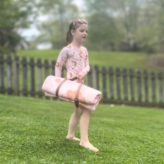 Young girl holding Blush Meadow Quilted Water Resistant Beach Mat on grass with a wooden fence in the background