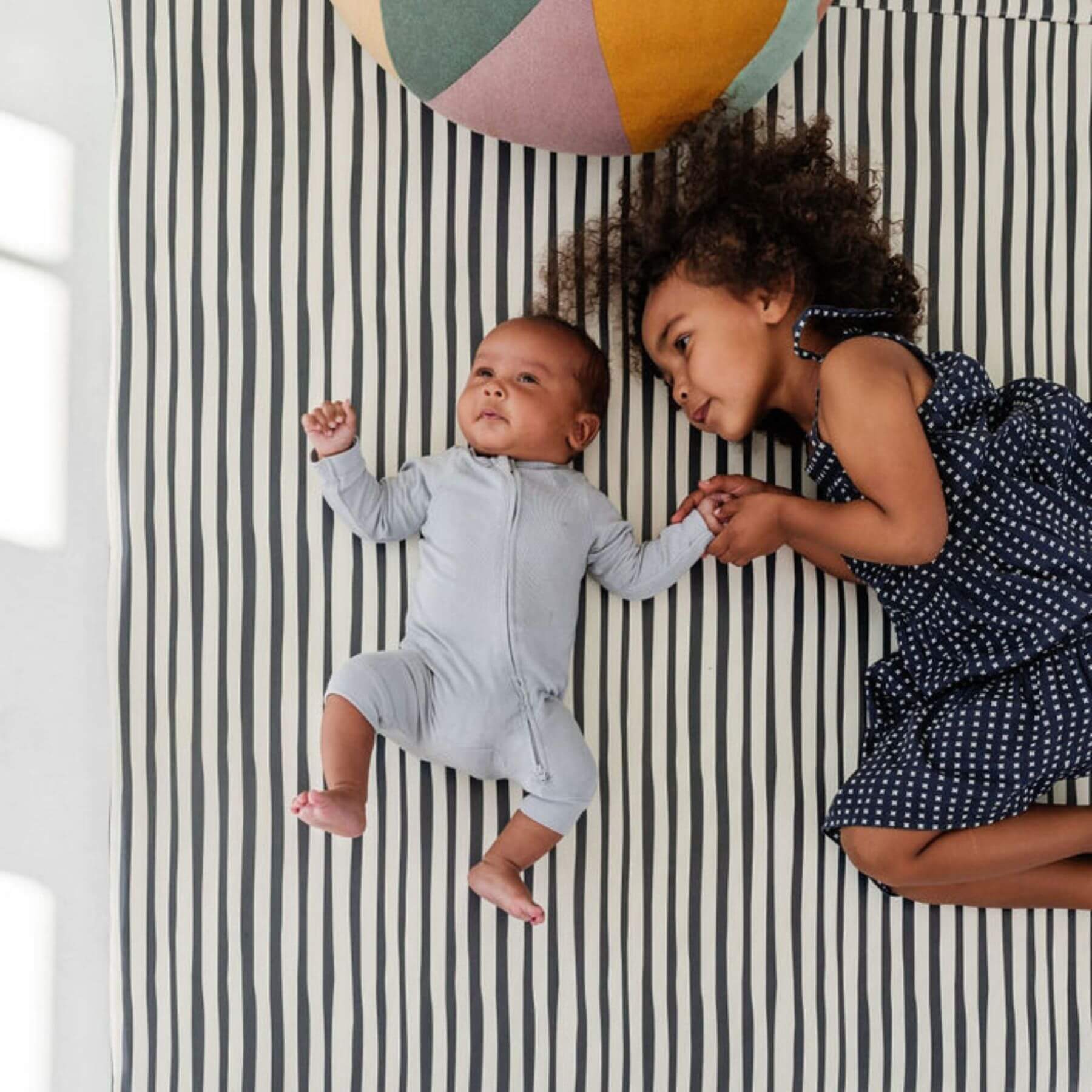 Two children lying on Toki Kids Bold Stripe Vegan Leather Mat with a colorful ball above them.