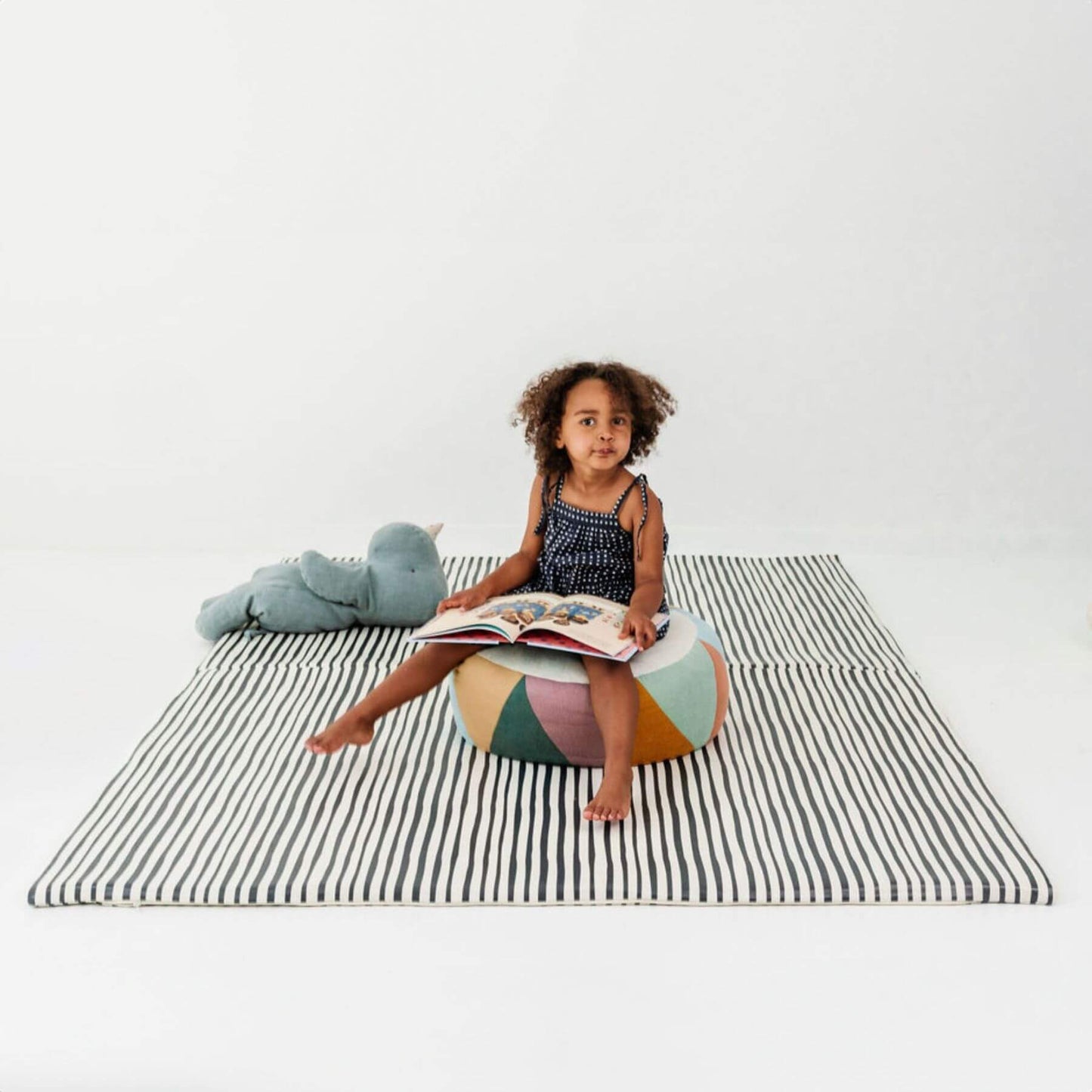 Child sitting on a colorful bean bag reading a book on Toki Kids Bold Stripe Vegan Leather Mat with a stuffed animal.