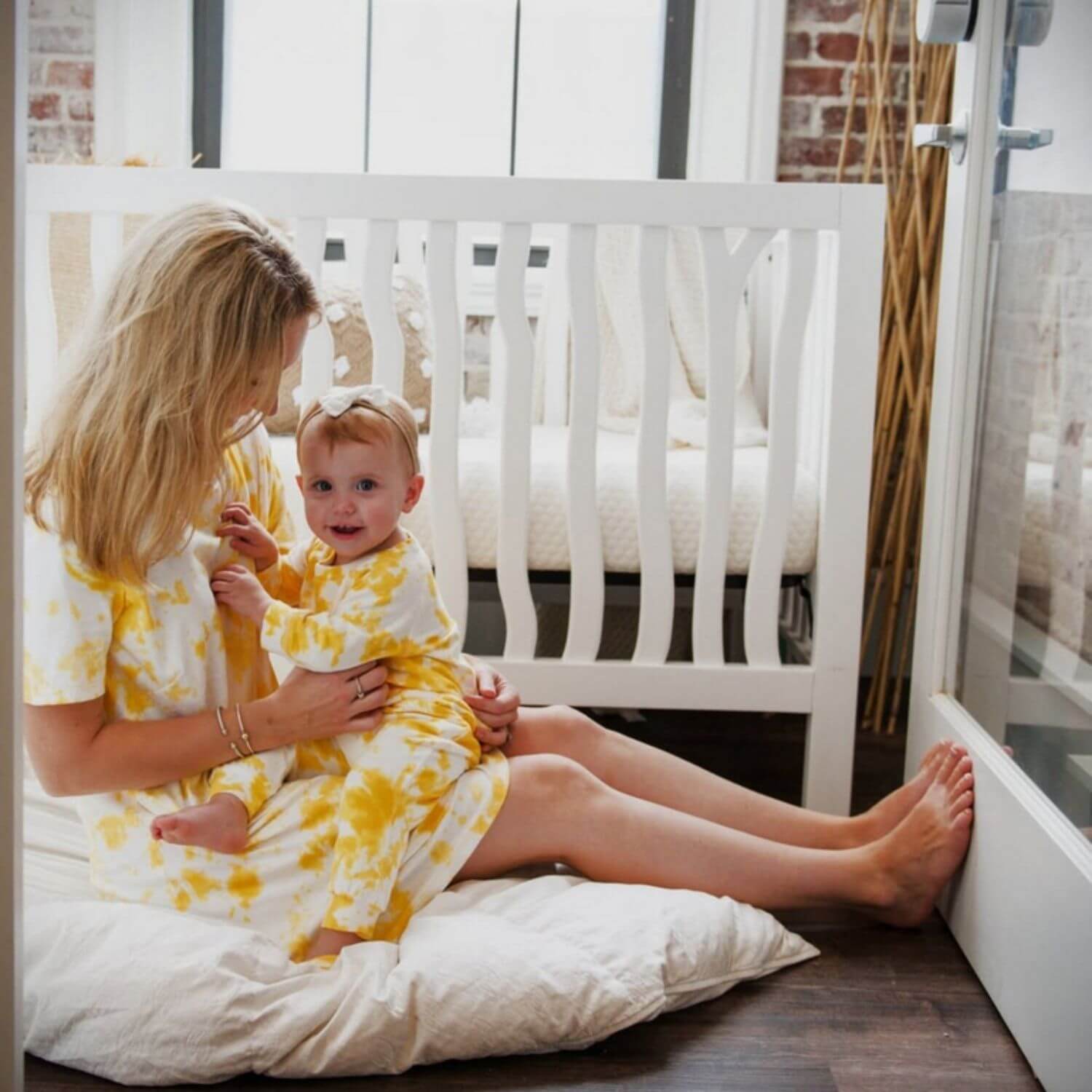 Woman and baby in matching yellow outfits sitting on a white pillow in a nursery.