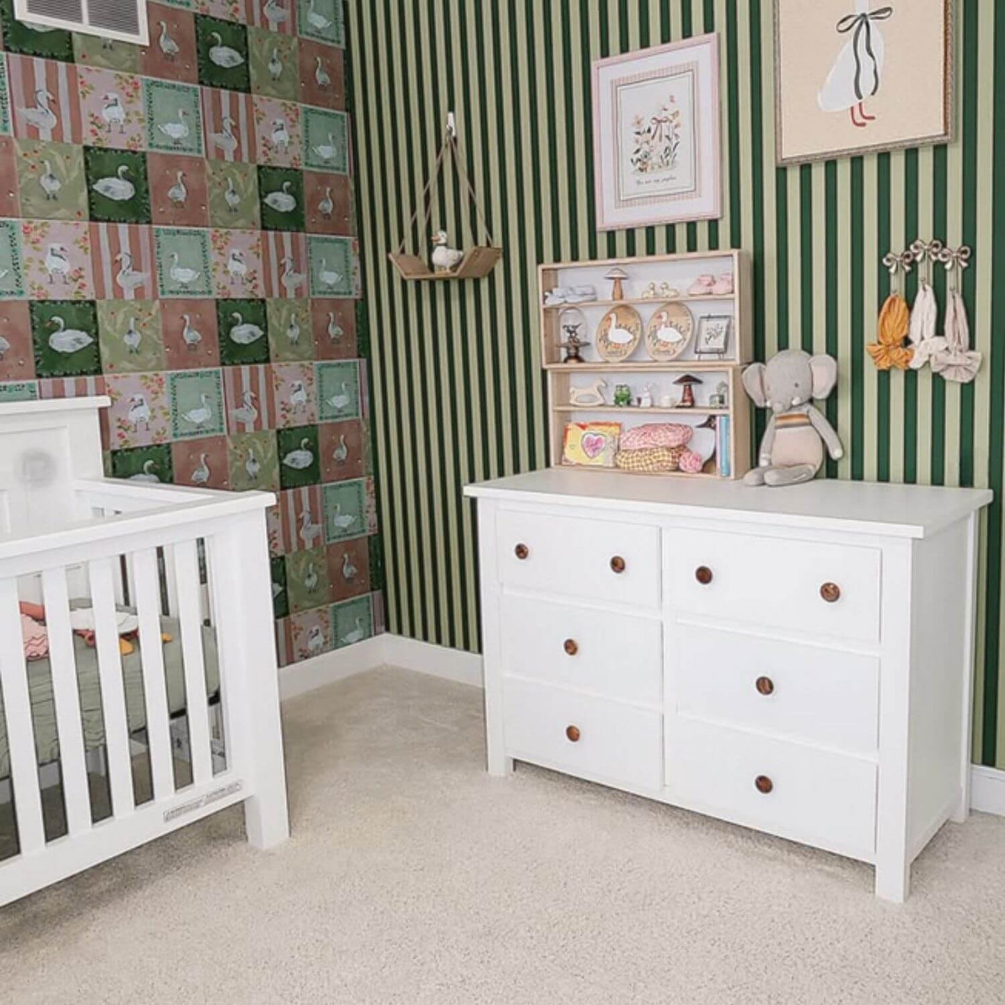 Nursery room with white dresser, crib, and decorative elements against a green and multicolored striped wall.