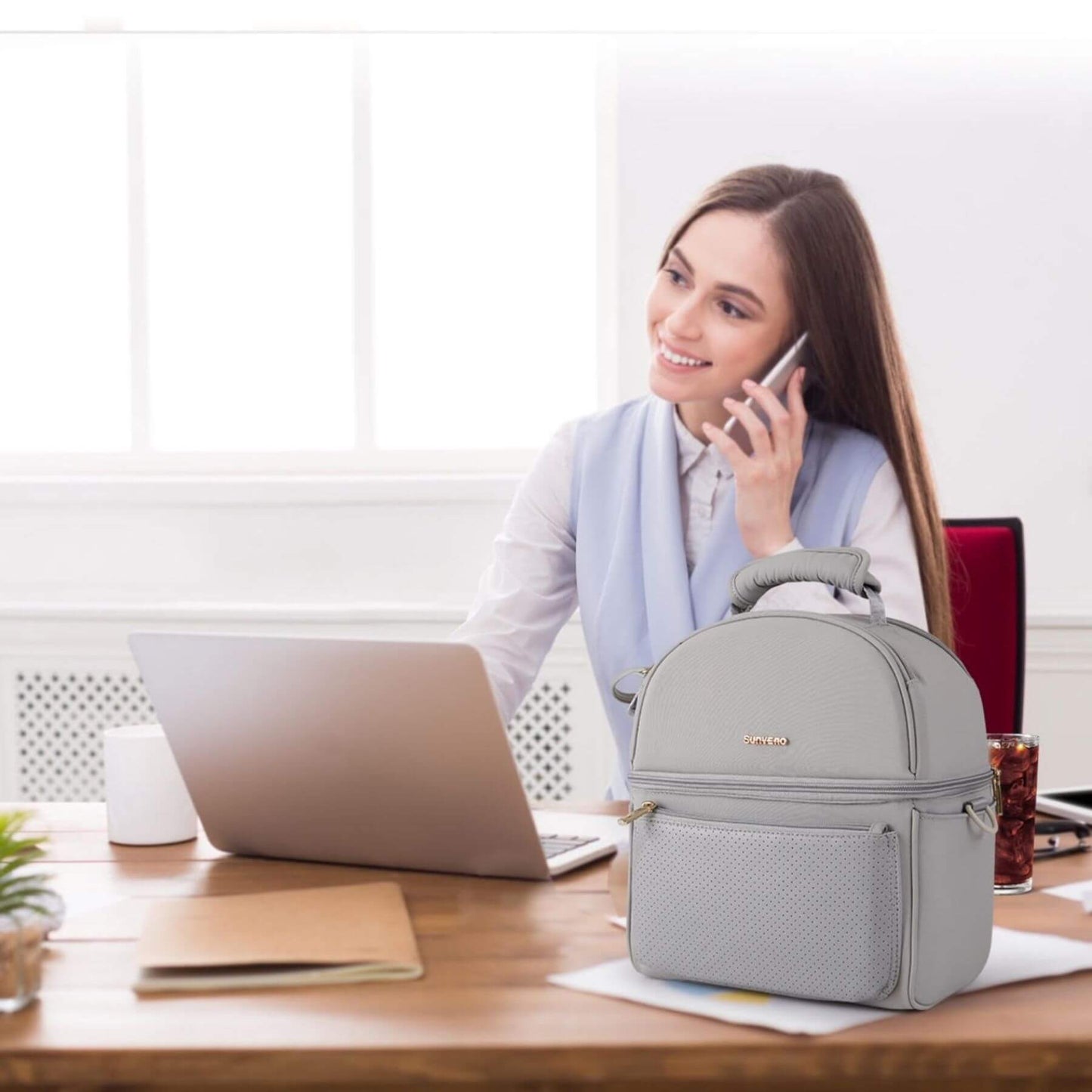 Woman sitting at a desk with a laptop and Sunveno Breastmilk Cooler Bag - Grey, talking on a phone.