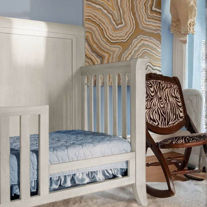 White crib with blue bedding in a room with a rocking chair and patterned wall.