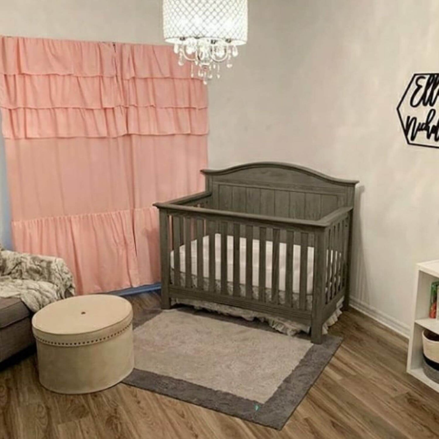 Nursery room with a gray crib, pink curtain, and round ottoman.