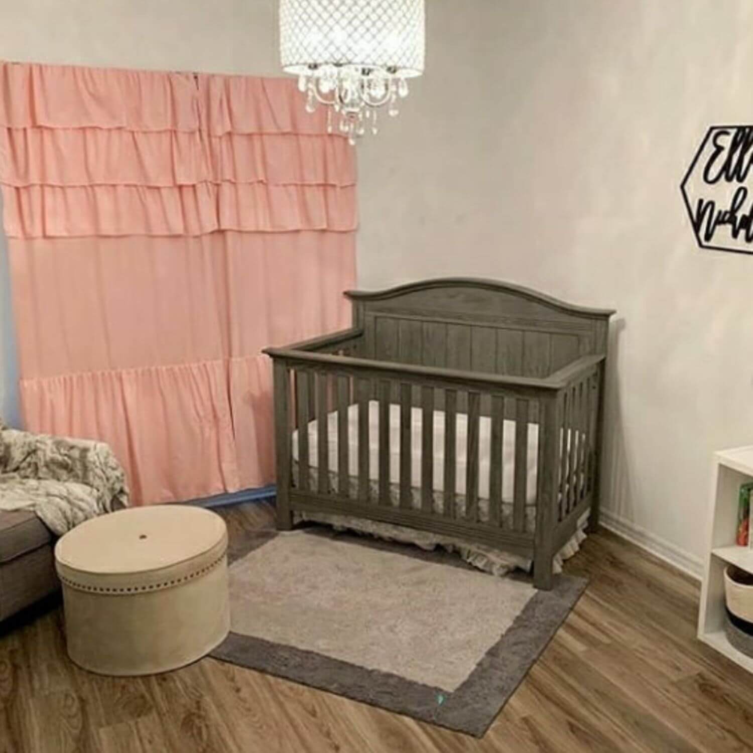Nursery room with a gray crib, pink curtain, and round ottoman.