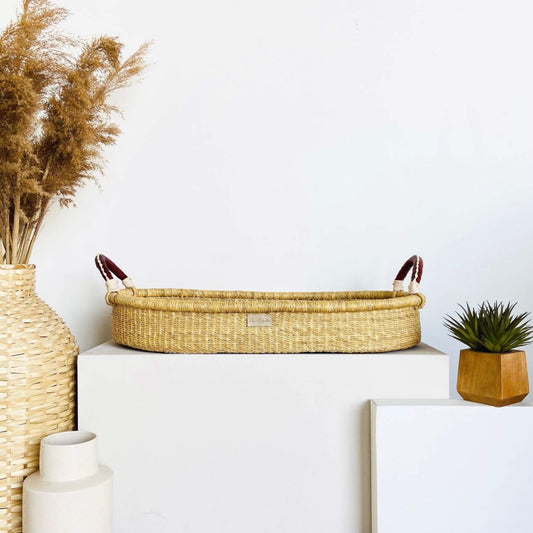 Plum+Sparrow Changing Basket Cognac + Cream Handle, vase with dried plants, and small plant on a white surface against a light blue wall.