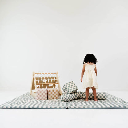 Child standing on Toki Kids Checker Matcha Organic Cotton Mat with pillows and a small wooden structure against a white background