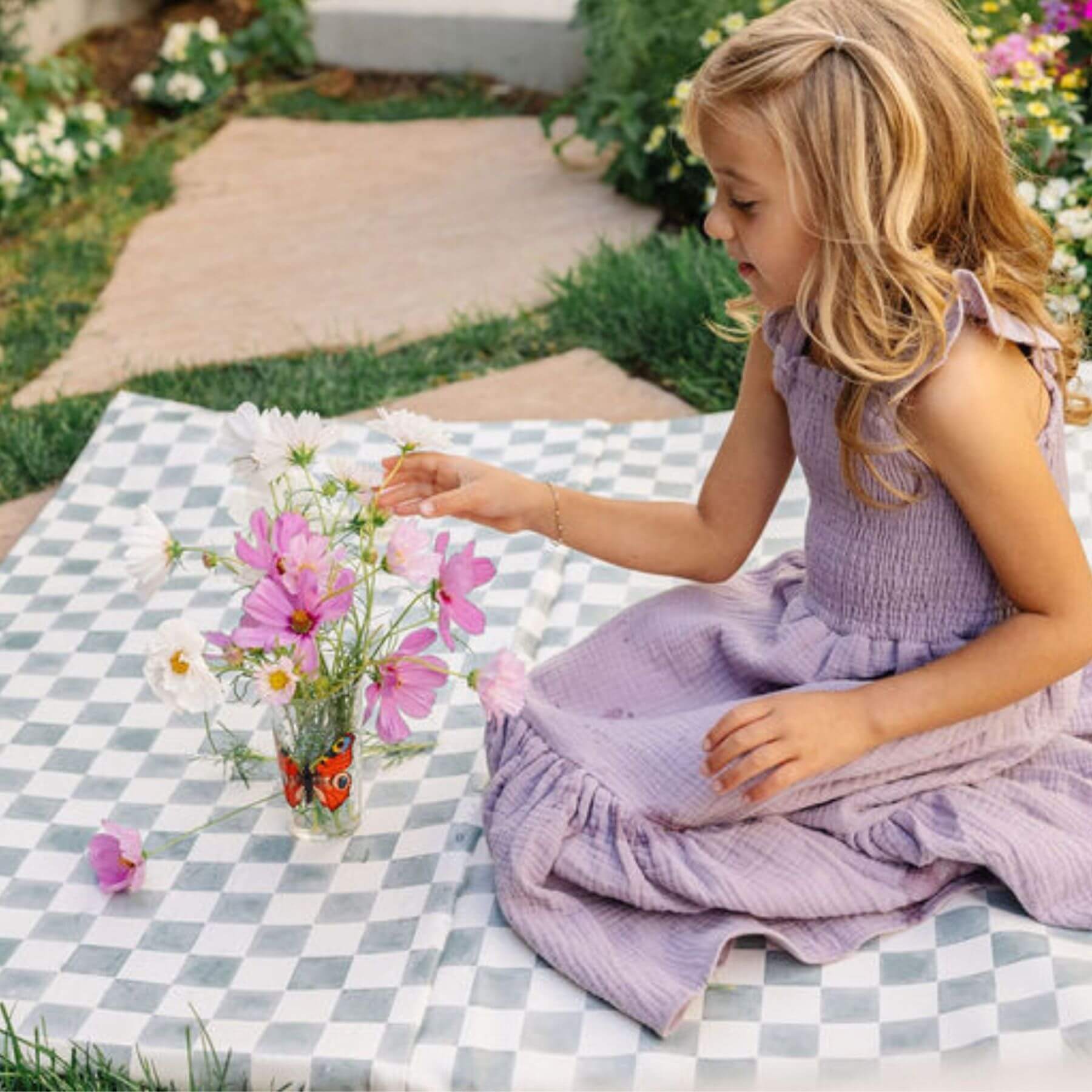 Young girl in a purple dress sitting on a checkered mat with flowers and a butterfly toy outdoors.