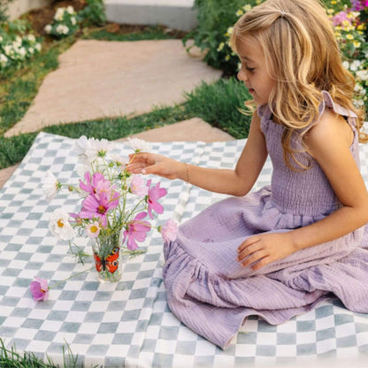 Young girl in a purple dress sitting on a checkered mat with flowers and a butterfly toy outdoors.