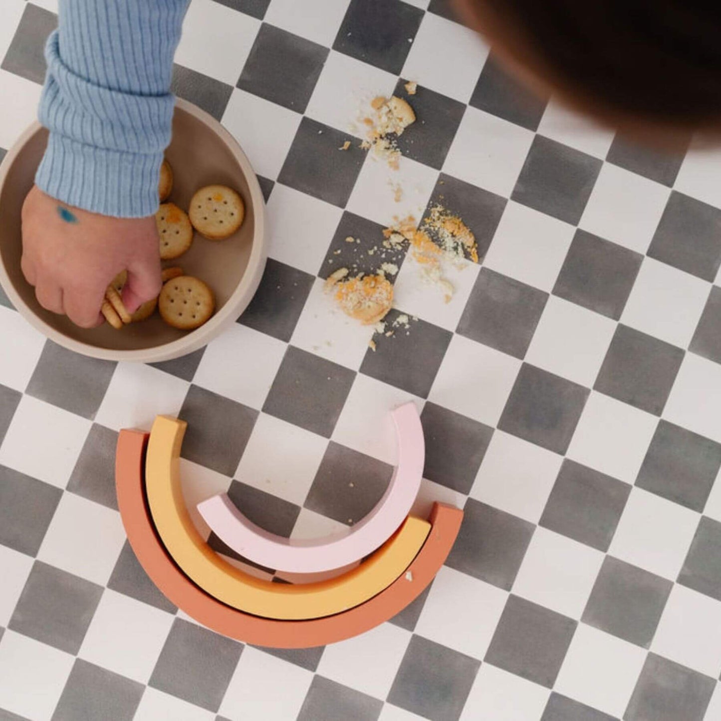 Colorful wooden toys on a checkered mat with a child's hand reaching for a bowl of crackers.