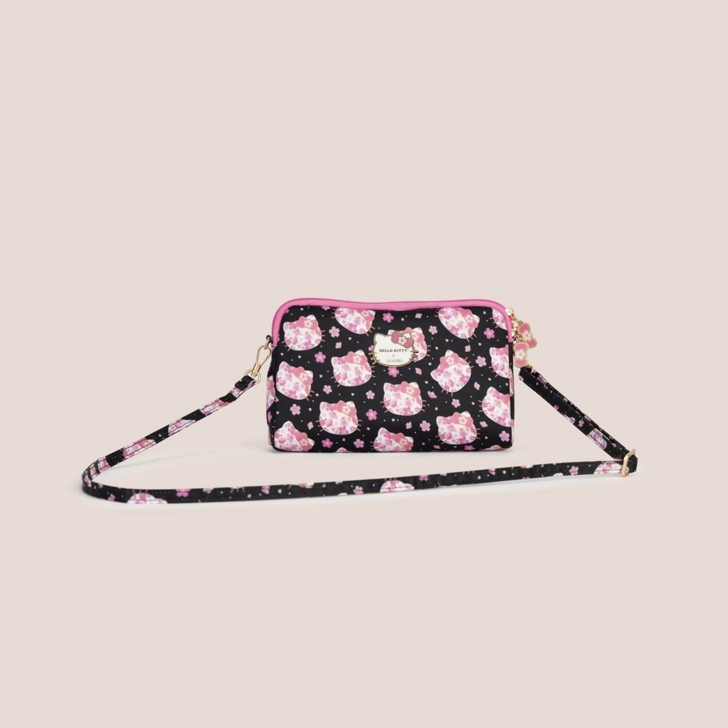A small crossbody bag with a black background and a pink Hello Kitty pattern, shown on a solid light beige background.