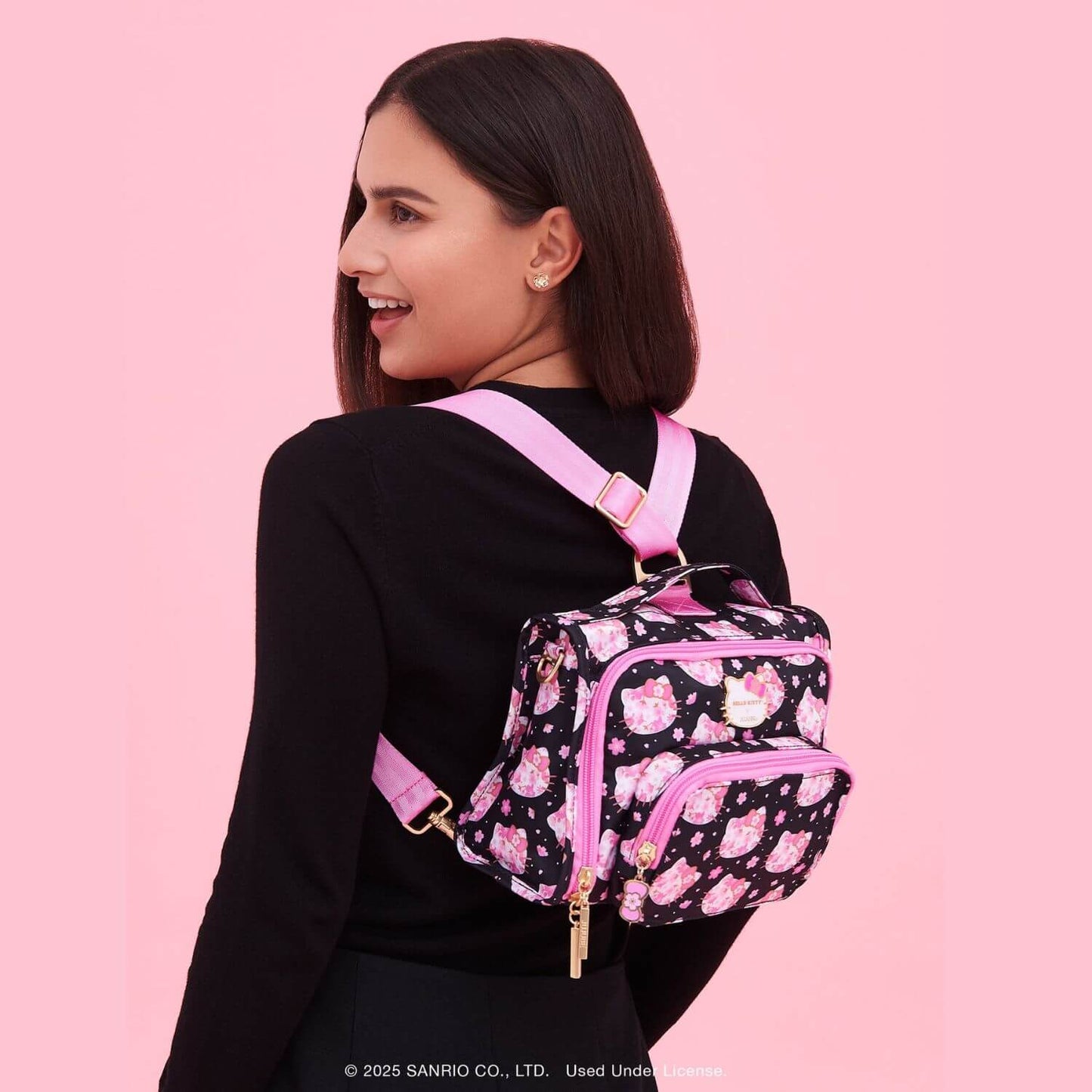 A woman wearing Hello Kitty® × JuJuBe Cherry Blossoms Bestie Small Messenger Handbag, set against a solid pink background.