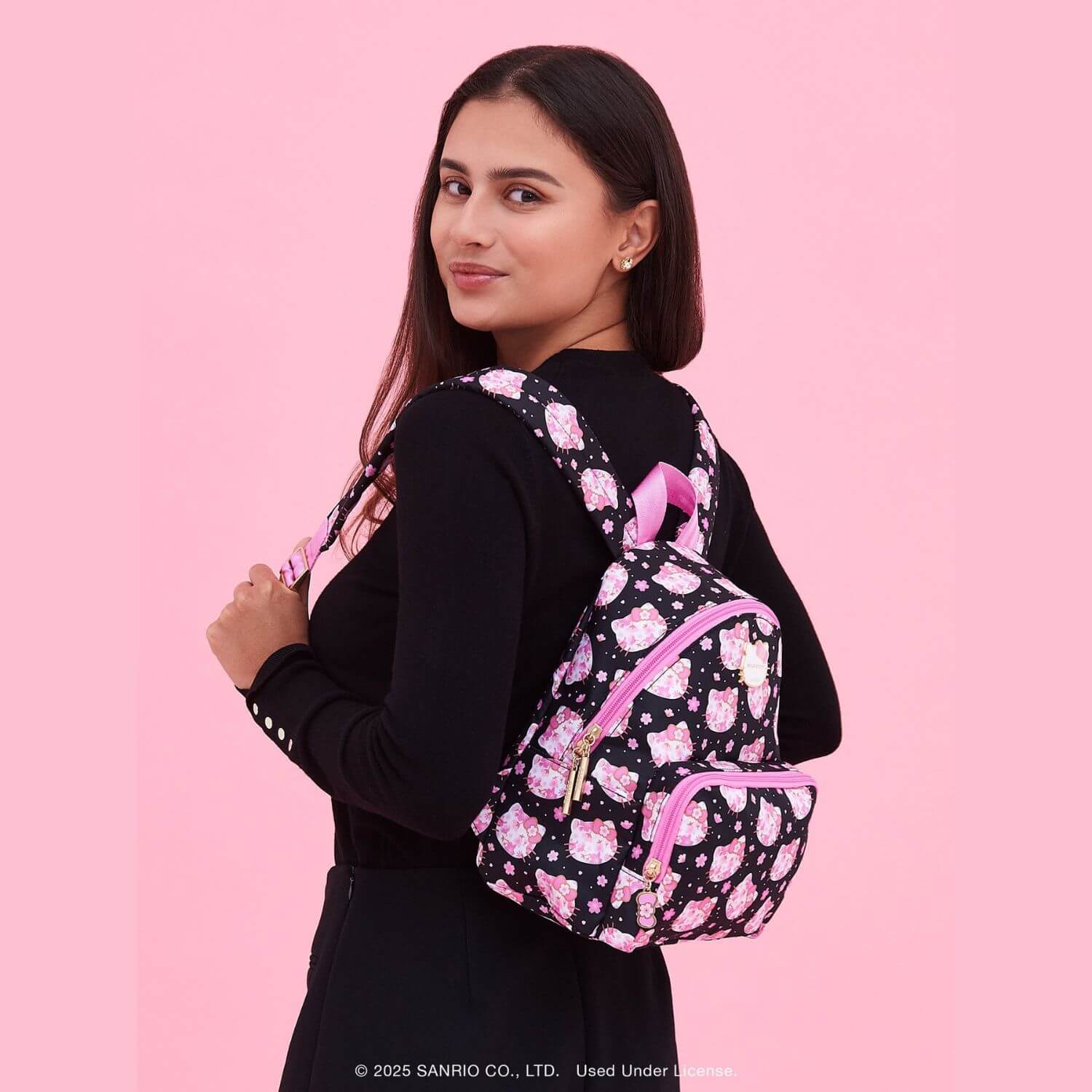 A woman wearing Hello Kitty® × JuJuBe Cherry Blossoms Everyday Mini Diaper Bag Backpack, set against a solid pink studio backdrop.