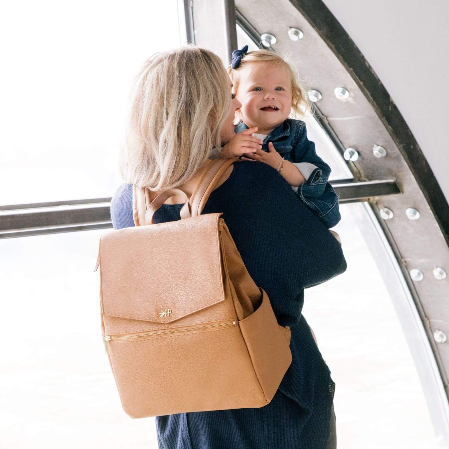 Woman holding a child wearing Butterscotch Classic Diaper Bag II in an indoor setting