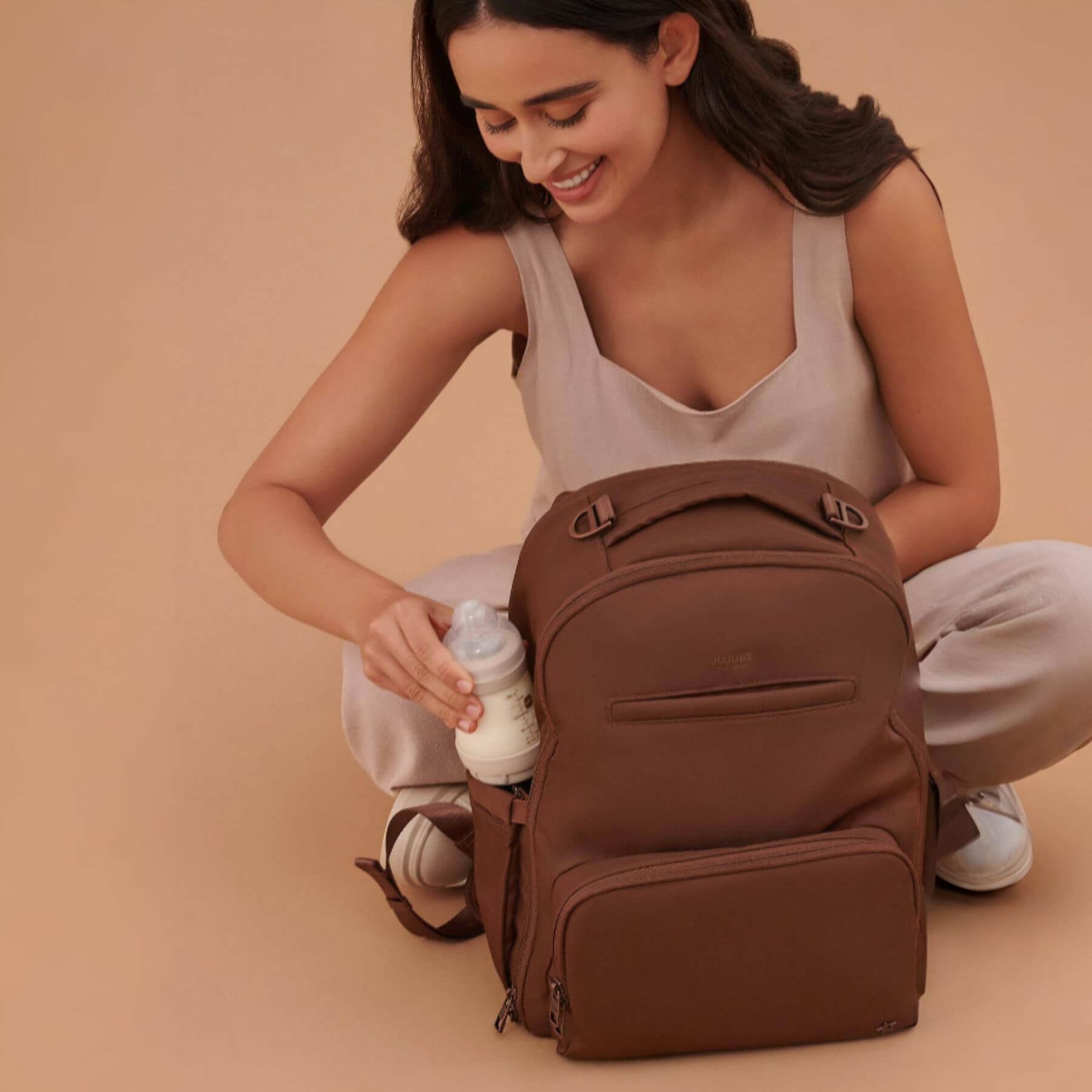 Woman sitting on the ground with JuJuBe Classic Diaper Backpack Espresso and a bottle, against a beige background