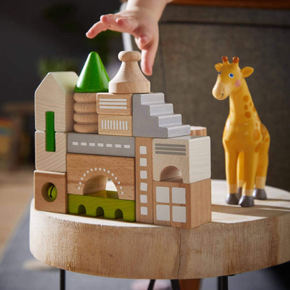 Wooden block set with a hand placing a green block, next to a toy giraffe on a wooden surface.