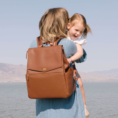 Woman carrying Cognac Classic Diaper Bag II with a child on her back, standing by a body of water.
