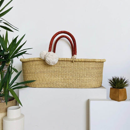 Plum+Sparrow Cognac+Cream Handle No Hood Moses Basket, displayed on a white pedestal against a white wall with potted plants nearby.