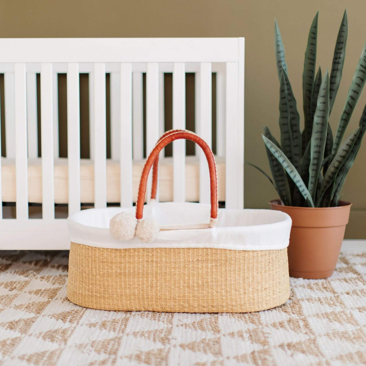 Plum+Sparrow Cognac+Cream Handle No Hood Moses Basket, styled in a nursery setting next to a white crib and a potted snake plant.