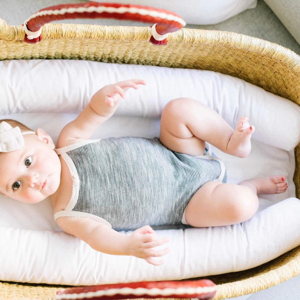 Baby lying in Plum+Sparrow Cognac+Cream Handle No Hood Moses Basket with white bedding and a striped blanket.