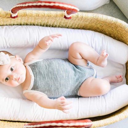 Baby lying in Plum+Sparrow Cognac+Cream Handle No Hood Moses Basket with white bedding and a striped blanket.