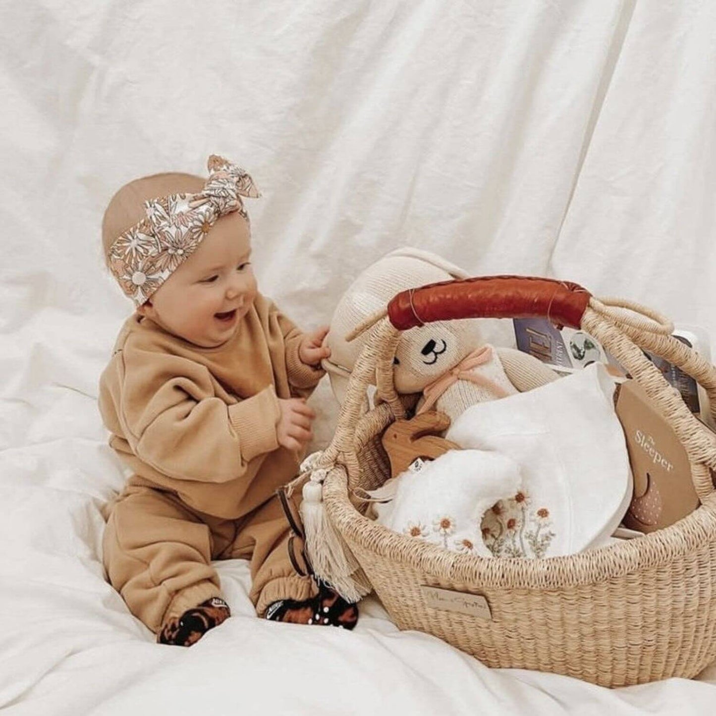 A baby in a tan outfit and silver bow headband sits beside Plum+Sparrow Cognac Handle Large Market Basket filled with baby accessories