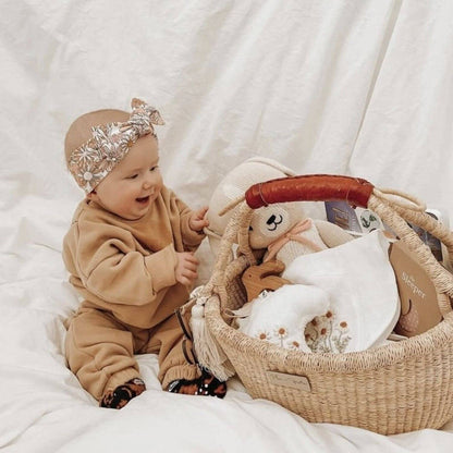 A baby in a tan outfit and silver bow headband sits beside Plum+Sparrow Cognac Handle Large Market Basket filled with baby accessories