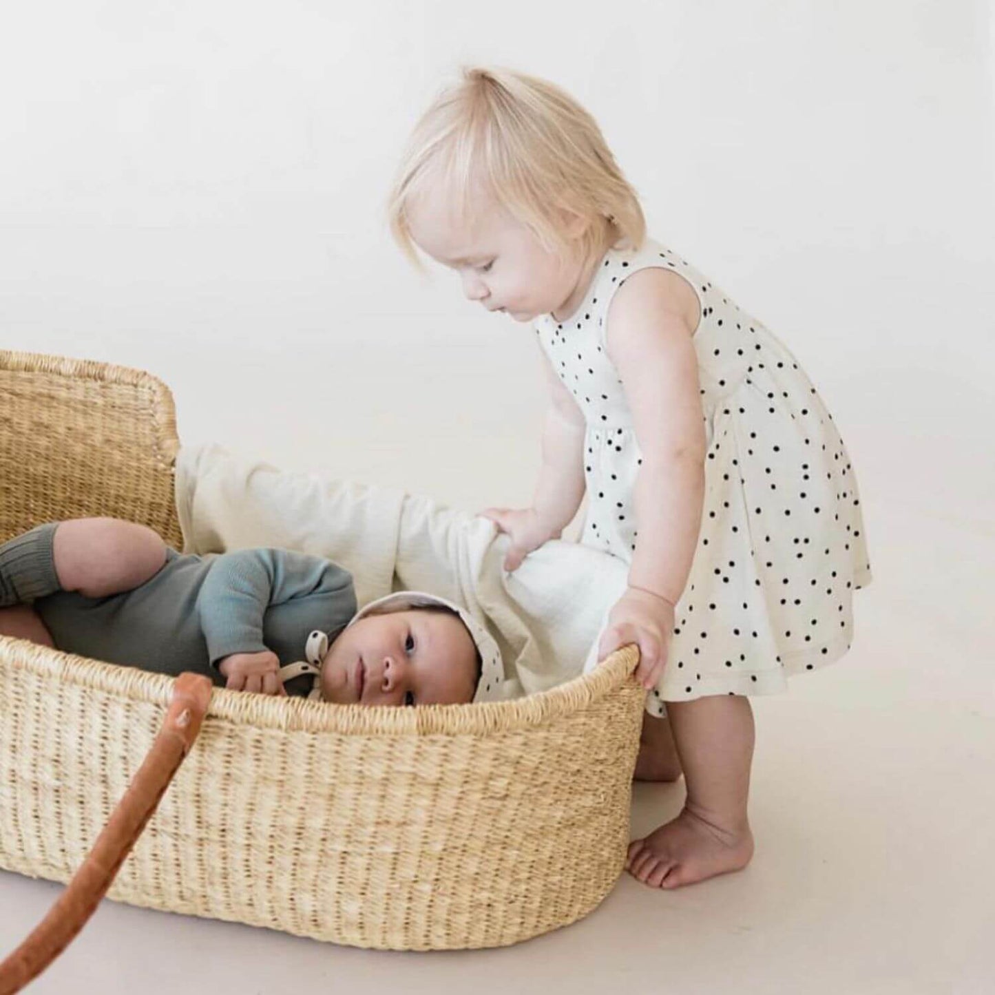 Baby lying in Plum+Sparrow Cognac Handle Moses Basket with a toddler standing next to the basket