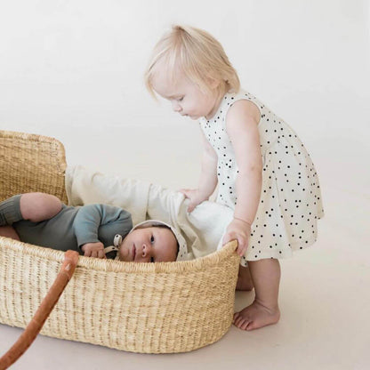 Baby lying in Plum+Sparrow Cognac Handle Moses Basket with a toddler standing next to the basket