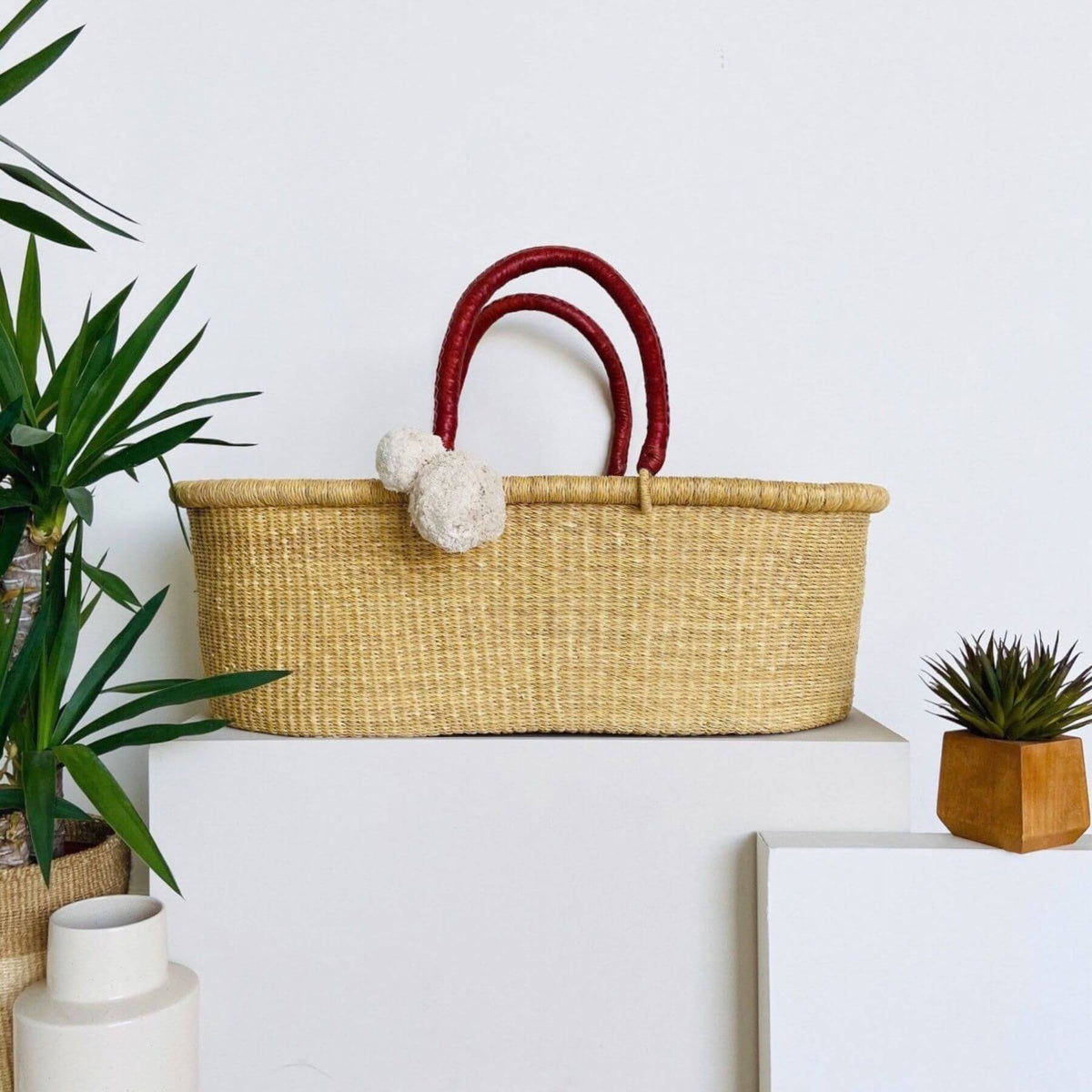 Plum+Sparrow Cognac Handle No Hood Moses Basket, displayed on a white pedestal with decorative plants in the background.