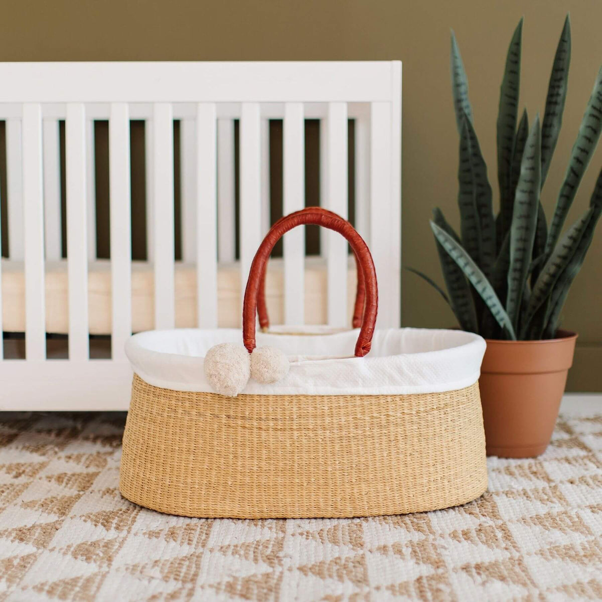 Plum+Sparrow Cognac Handle No Hood Moses Basket, styled on a patterned rug in front of a white crib and next to a potted snake plant.