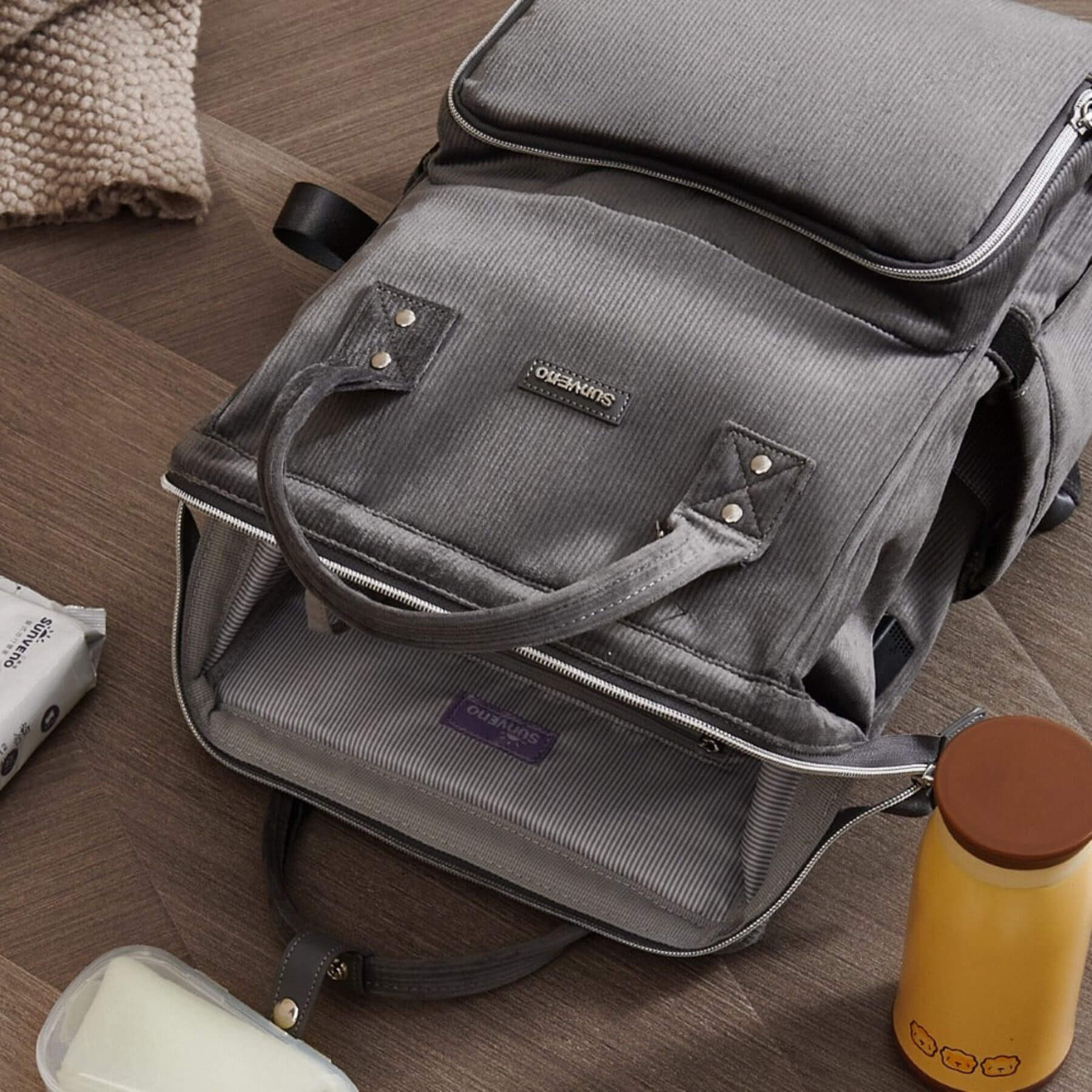 Corduroy Diaper Backpack Grey on a wooden floor with a yellow thermos and white container nearby.