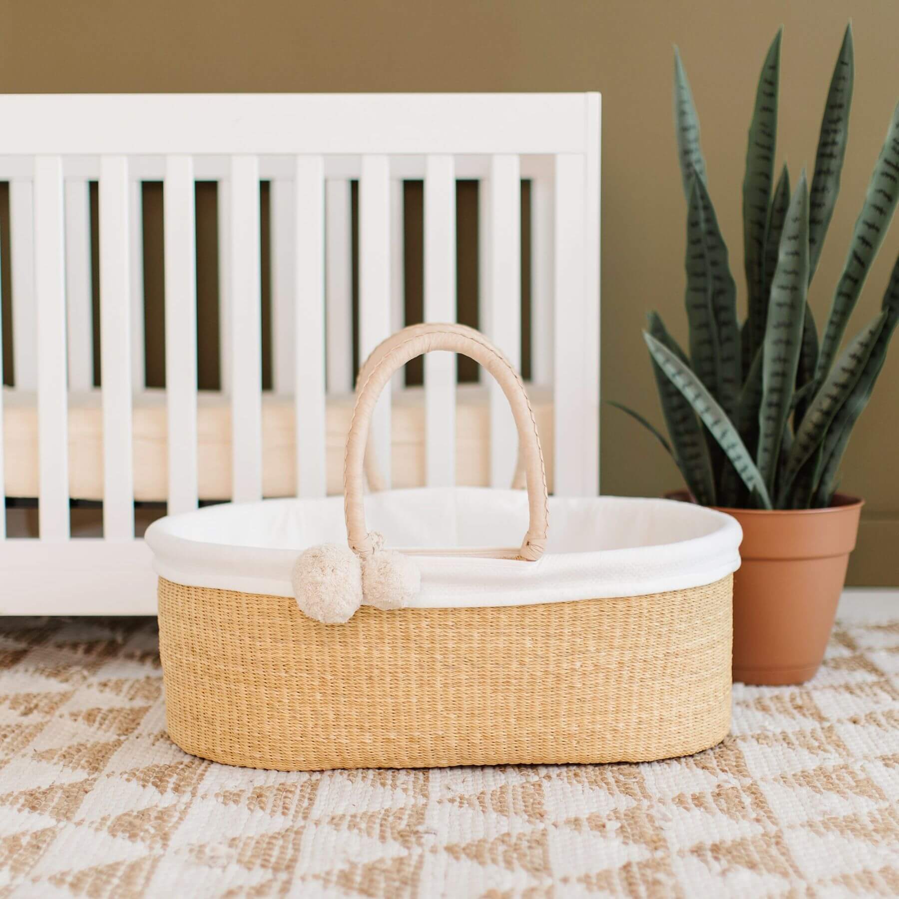 Plum+Sparrow Cream Handle No Hood Moses Basket on a checkered floor in front of a crib.