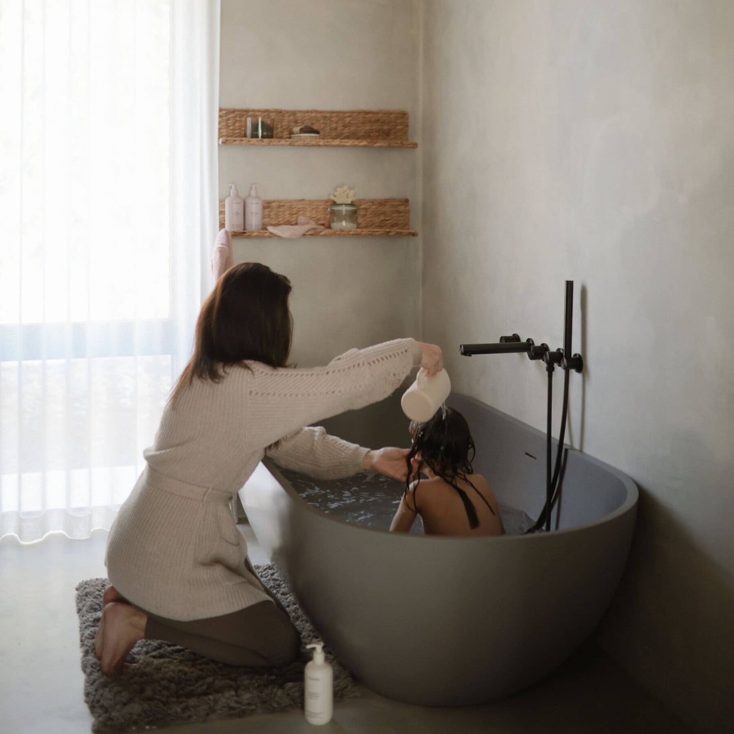 Woman in a beige sweater and shorts kneeling beside a bathtub with another person inside, in a bathroom setting.