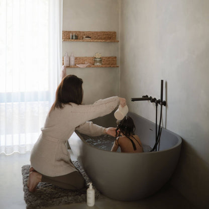 Woman in a beige sweater and shorts kneeling beside a bathtub with another person inside, in a bathroom setting.