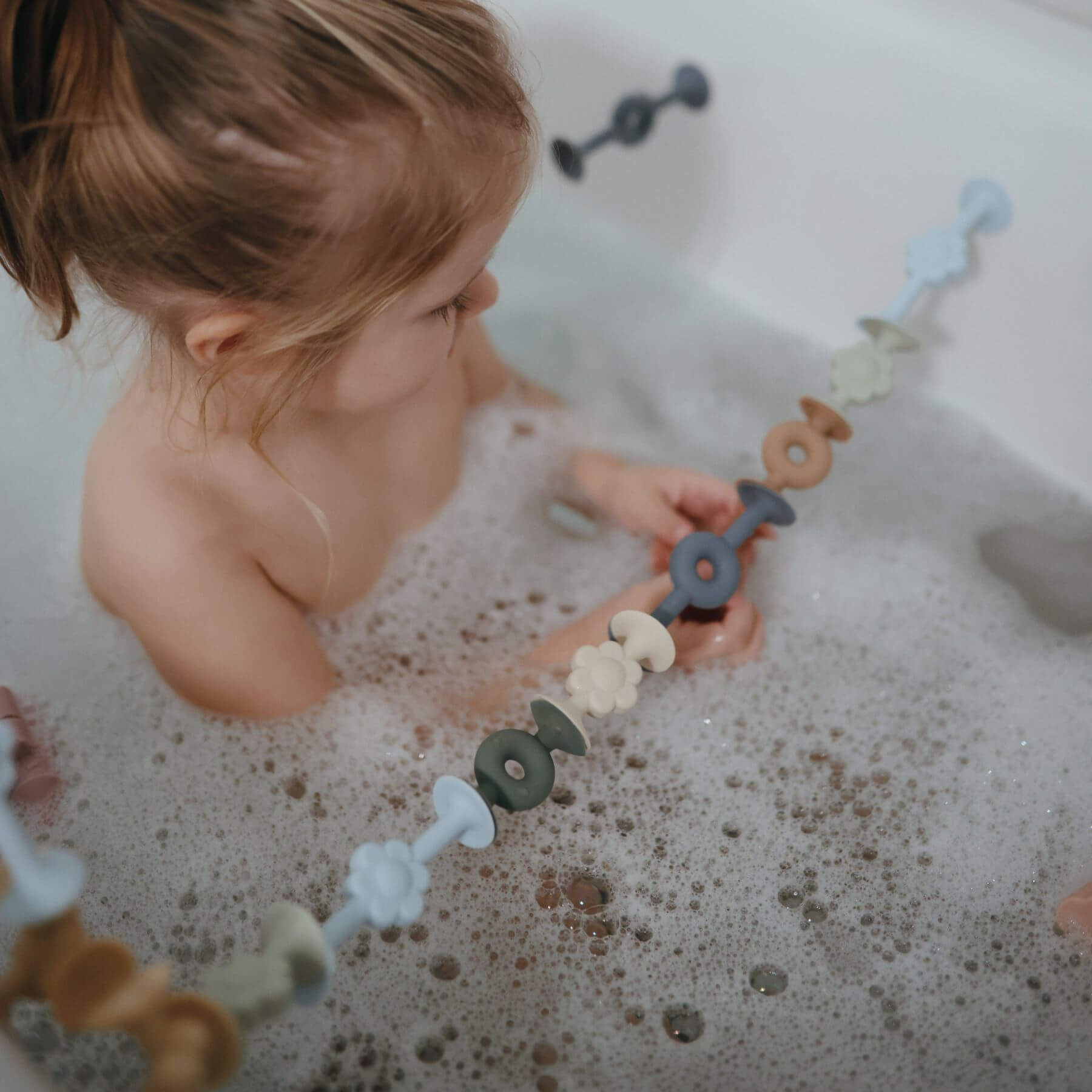 Child playing with Mushie Donut Silicone Suction Builders in a bathtub filled with bubbles