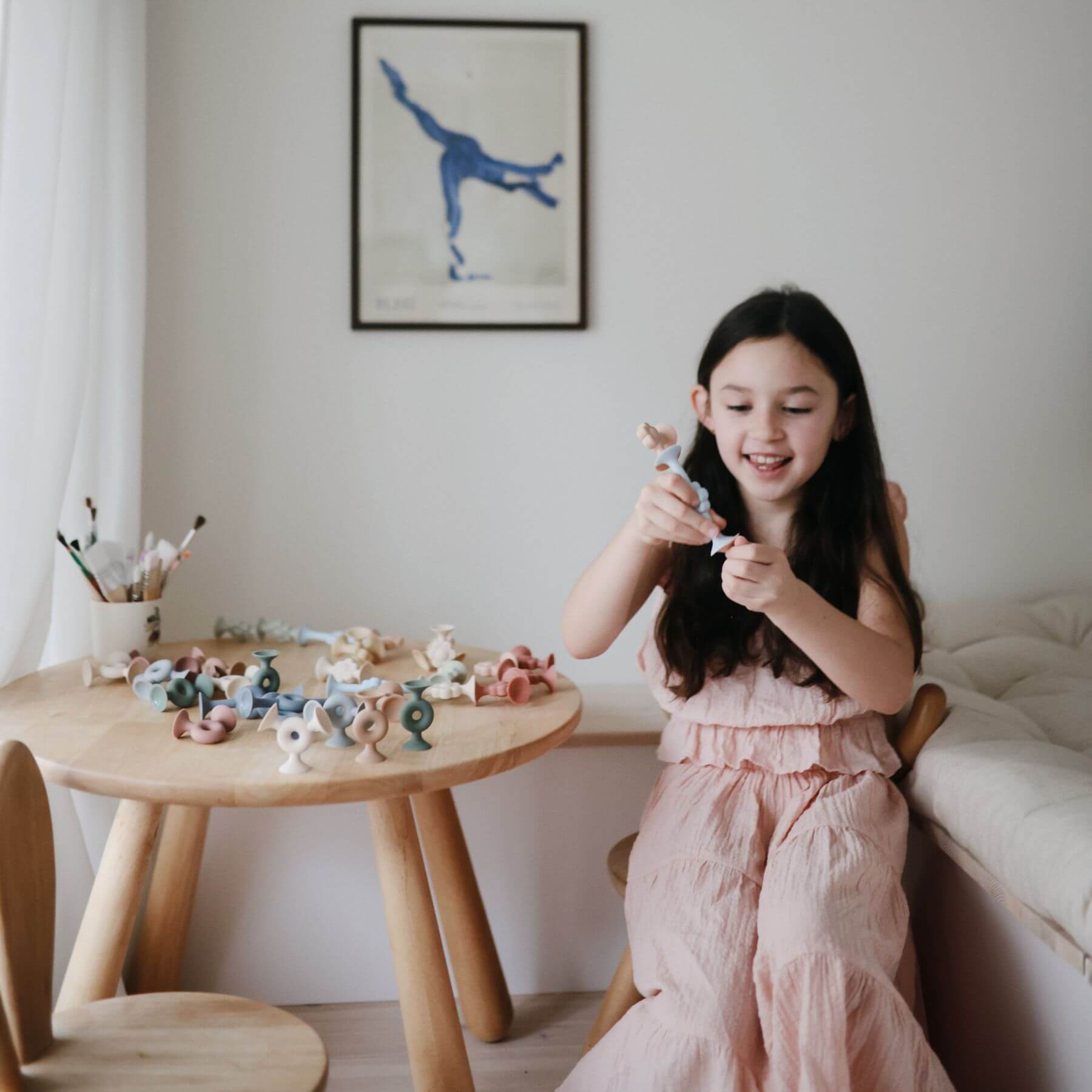 Young girl in a pink dress playing with Mushie Donut Silicone Suction Builders in a room with a wooden table and framed artwork on the wall.