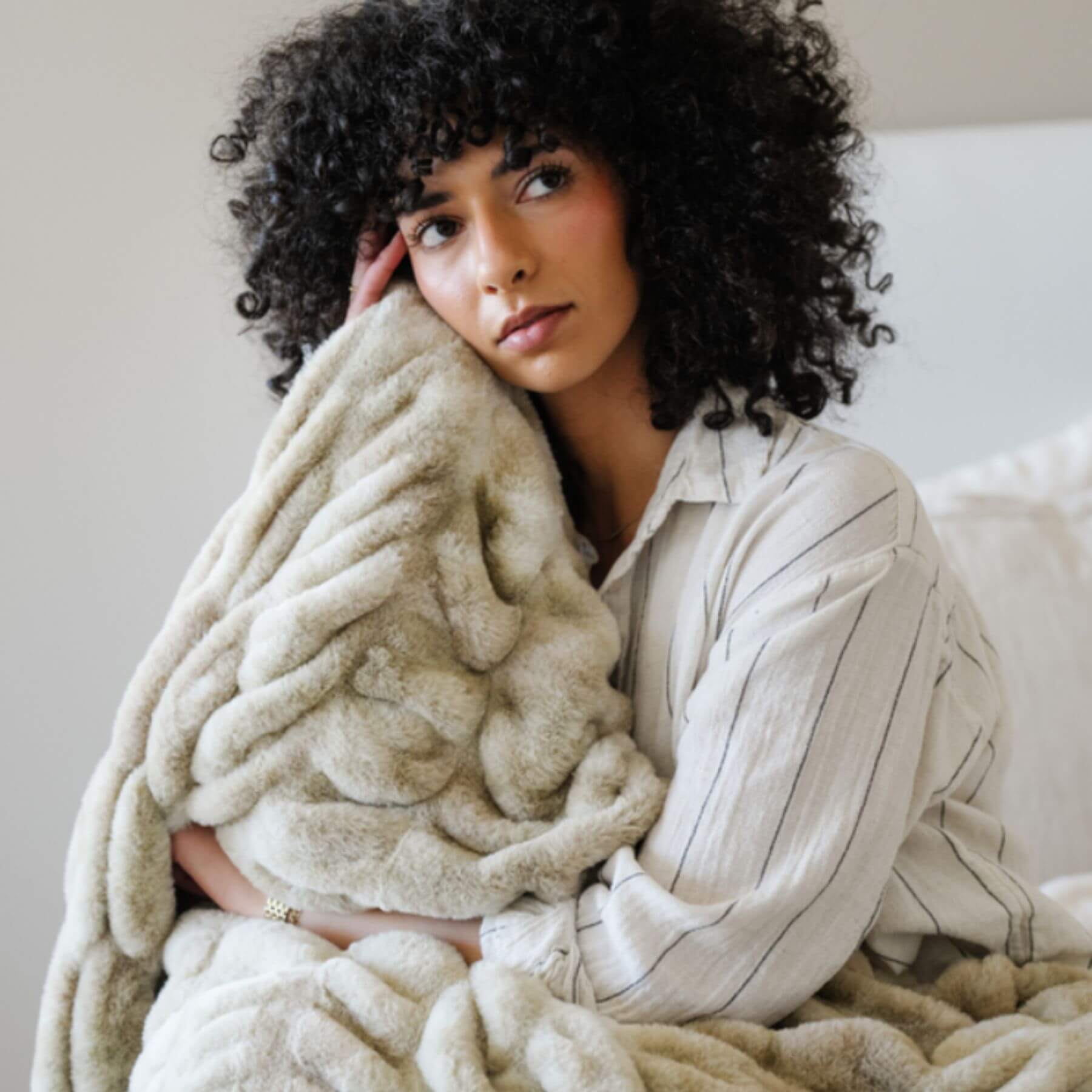 Woman wrapped in Saranoni Double Ruched Faux Fur Throw Blankets Sand, sitting on a couch in a cozy indoor setting.